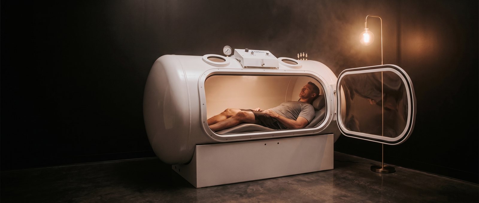 Man resting inside a hard-shell hyperbaric oxygen chamber with the door open, dark studio