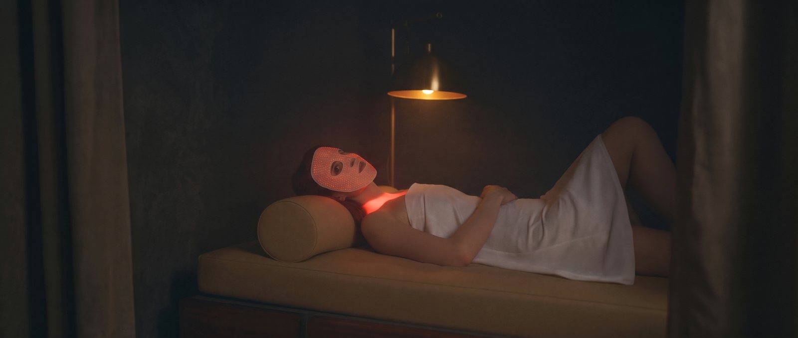 Woman lying under a lamp wearing a lit red LED face mask in a dark studio
