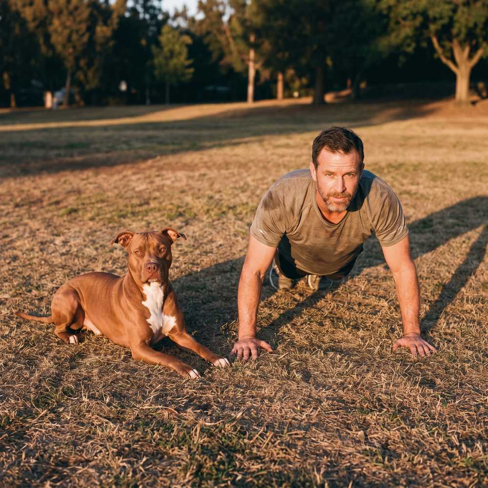 A man holding a push-up on a park lawn with a pit bull lying beside him